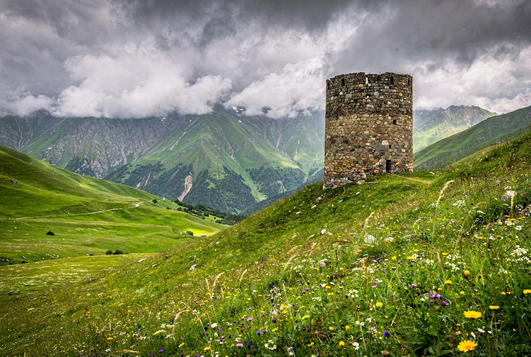 Lush green alpine valley with a stone watchtower on a hillside, wildflowers in foreground, misty Caucasus peaks behind