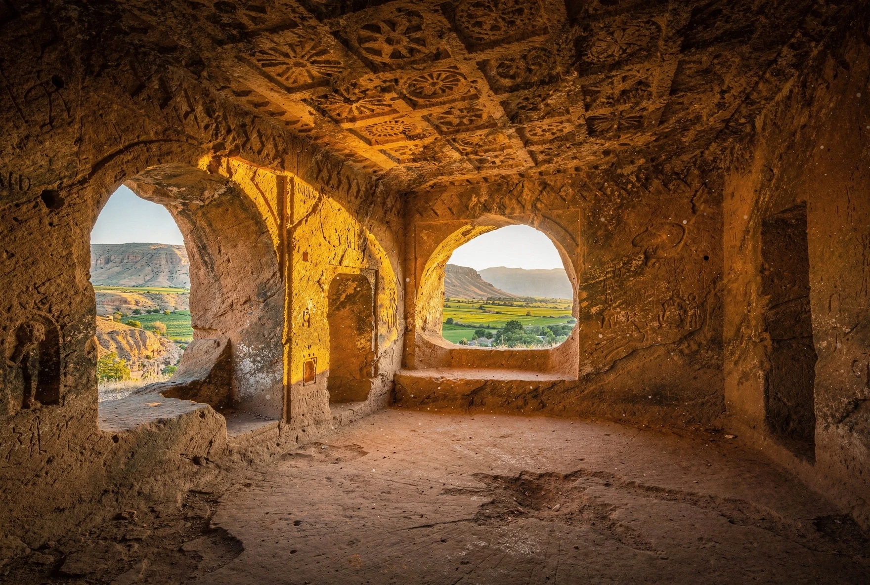 Rock-carved chambers with coffered ceiling details in sandstone, looking out over a green valley through cave openings