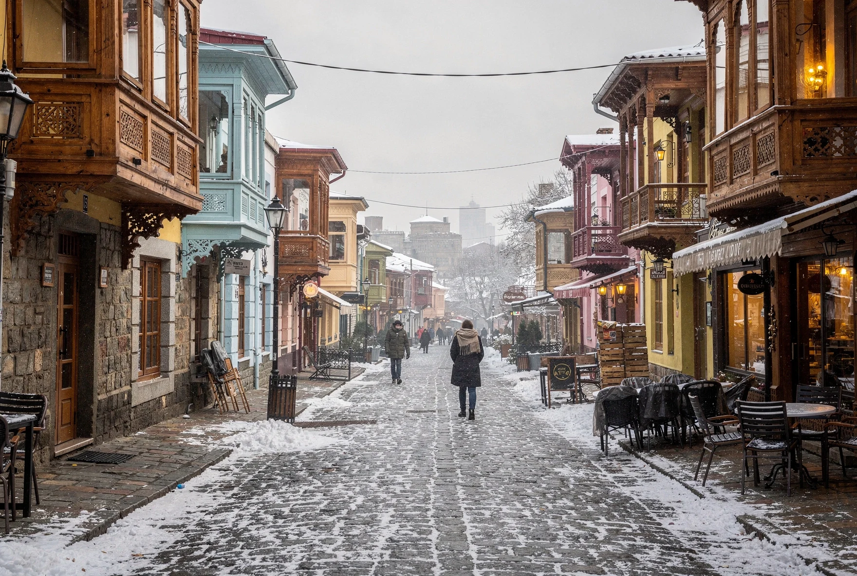 Tbilisi Old Town street with traditional wooden balconies covered in light snow, warm window light, people walking in winter coats